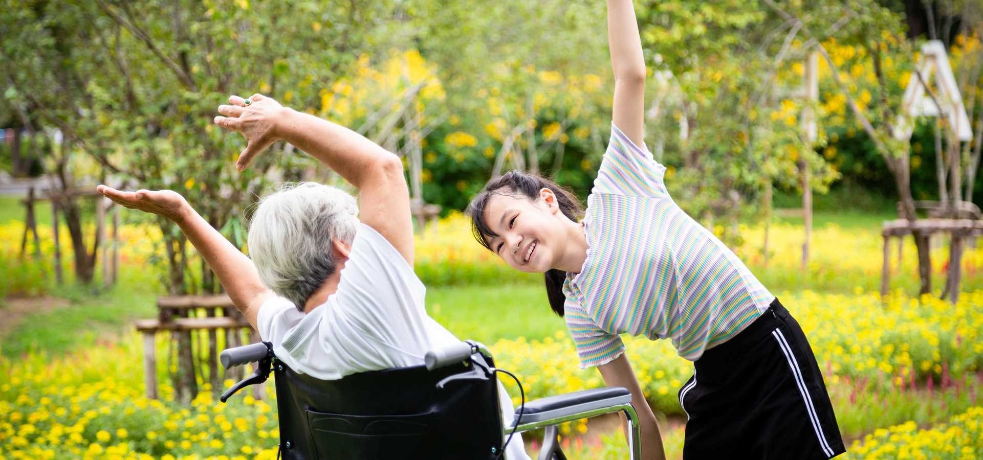 Ältere Dame im Rollstuhl und junge Frau machen Bewegungsübungen im Park