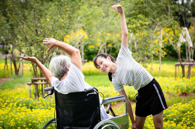 Ältere Dame im Rollstuhl und junge Frau machen Bewegungsübungen im Park